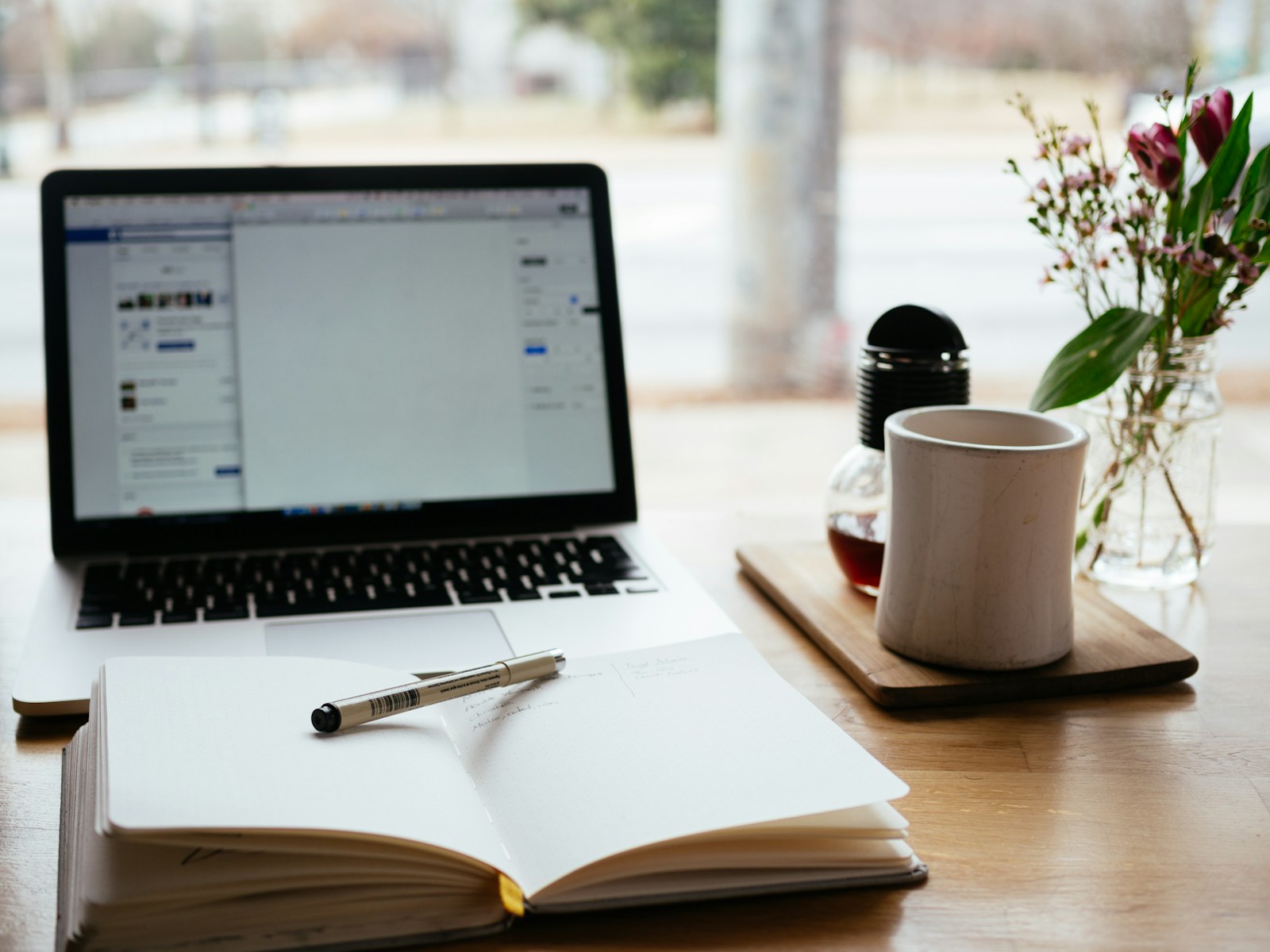 An open notebook and laptop on a wooden table with coffee and flowers